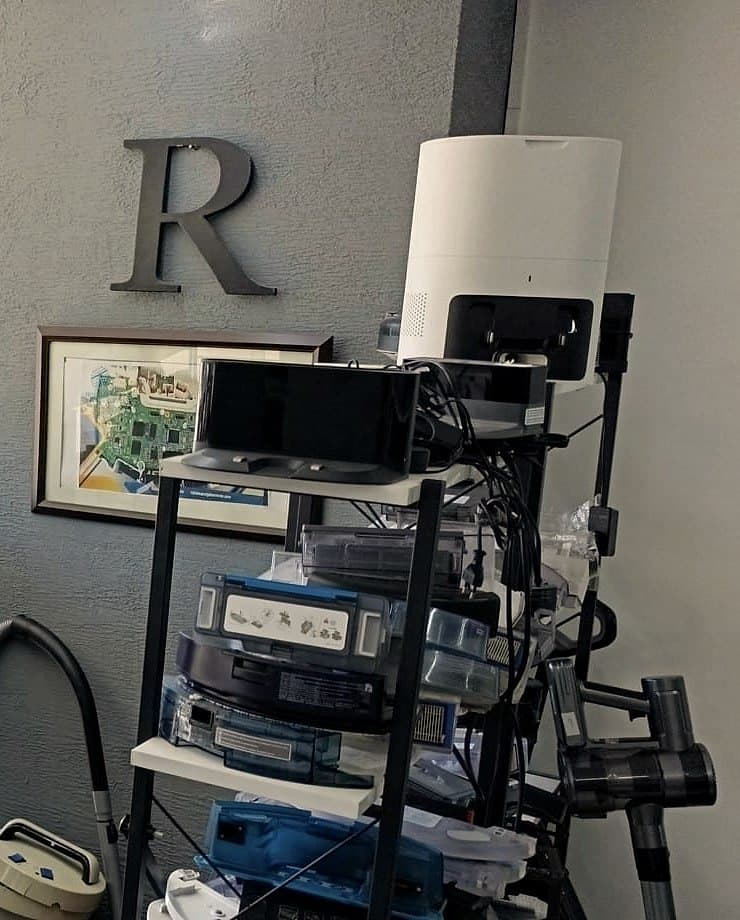 Atölye duvarında R harfi ve çerçeveli elektronik kart çizimi; önünde istasyon ve hazne parçalarıyla dolu raf