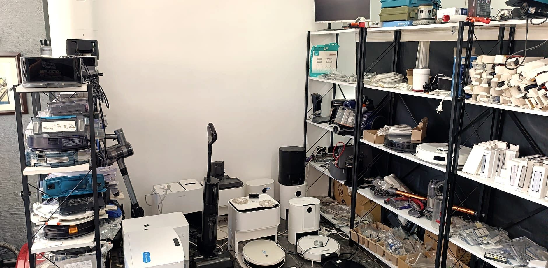 Atölye zemininde robot süpürgeler ve şarj istasyonları, arkada parçaların dizildiği raflar