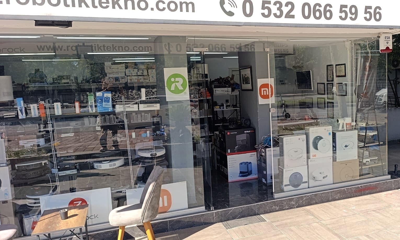 Dükkân vitrini; camın arkasında robot süpürge kutuları ve raflar, üstte telefon numaralı tabela